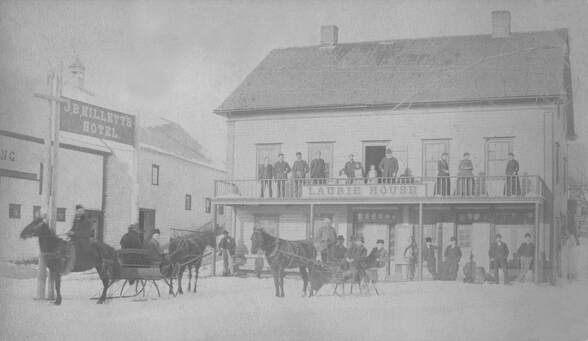 Very early image of the Town Hall building. On the left is a barn with a sign reading JB Millet's Hotel. The Town Hall building has a sign reading Laurie House. There are many people posing on the porches and some horses pulling buggies in the fore ground.